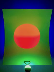 Photo of lighted wall sculpture of orange circle of light in green field of light in a blue field of light on wall behind.